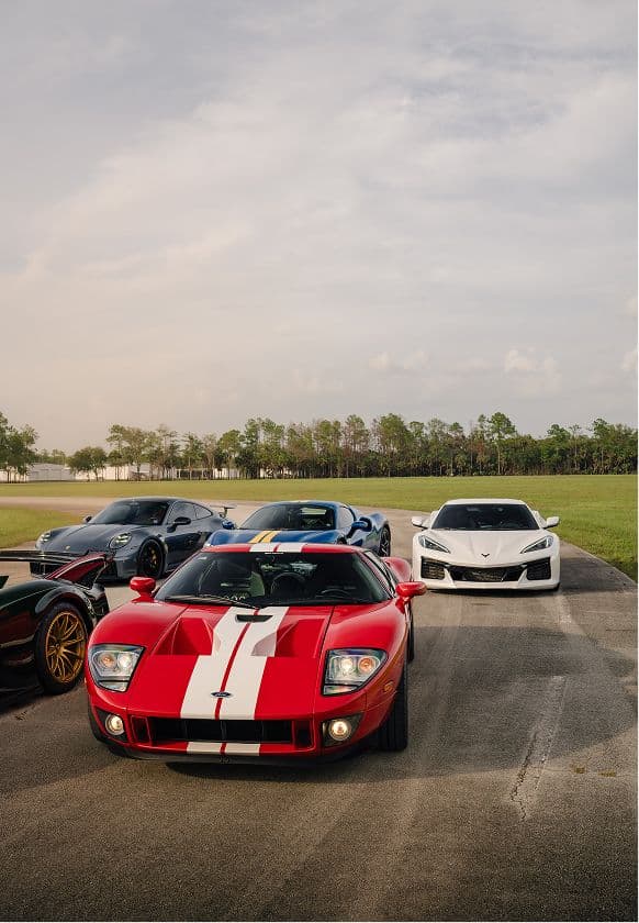 A red Ford GT with racing stripes leading a group of sports cars parked on the property, pine woodland behind.
