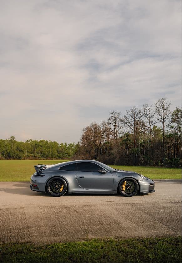 A grey Porsche 911 parked on the apron of the former proving ground, backed by Florida pine woodland.